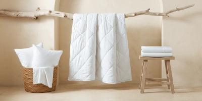 A white quilted comforter or duvet hangs from a rustic wooden branch in a warm, neutral-toned room. To the left is a woven basket holding folded white pillows and a sheet, and to the right is a small wooden stool with a folded stack of white towels.