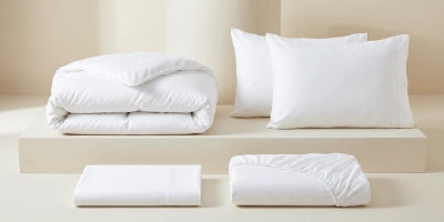 White bedding set including a folded duvet, two pillows, a folded sheet, and a folded fitted sheet, arranged on a minimalist beige platform against a warm cream background.