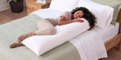 A woman lies on a bed using a long white body pillow to support her side, wearing beige pajamas in a brightly lit bedroom.
