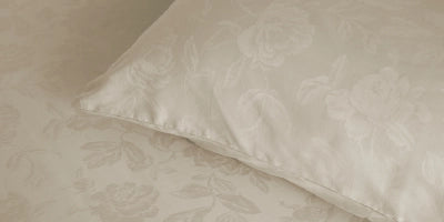 A close-up of a cream-colored pillowcase with a subtle tonal floral damask pattern, resting on matching patterned bedding.